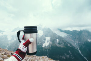 Closeup photo of thermos mug with tea in traveler's hand over out of focus mountains view with snow, tourizm in cold season