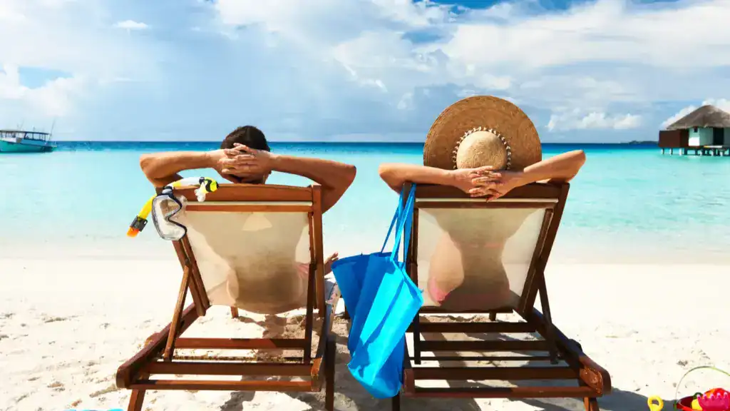 Couple relaxing on lounge chairs under the sun, facing turquoise ocean water with an overwater bungalow in the background.