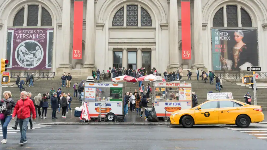 Crowds of tourists and food carts in front of the Metropolitan Museum of Art in New York City with a yellow taxi passing by.
