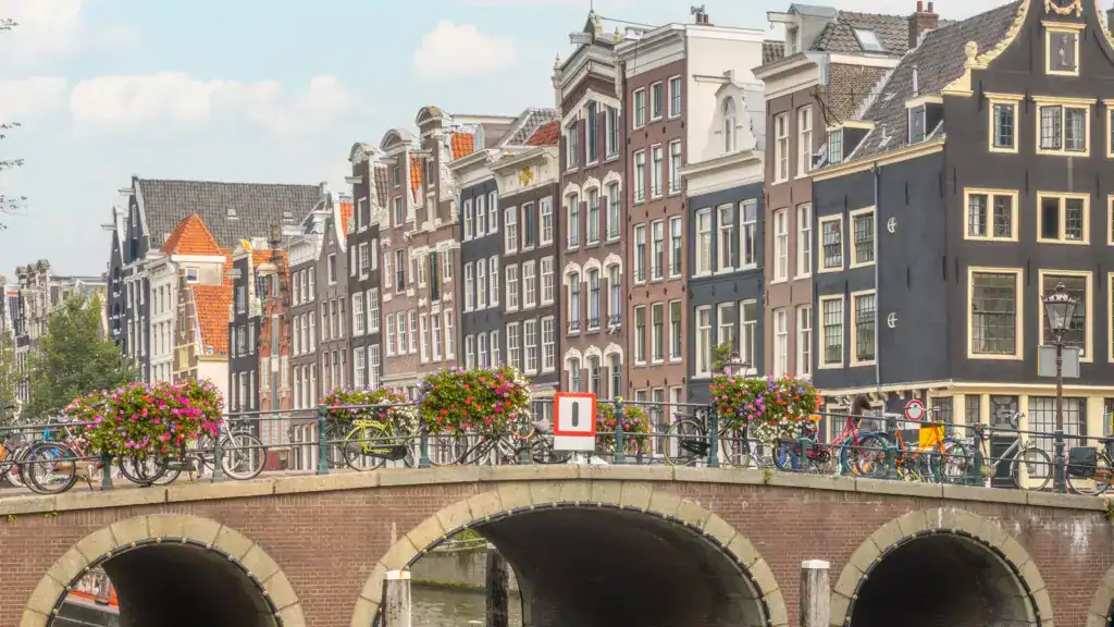 Historic stone bridge over an Amsterdam canal with bicycles, flower baskets, and traditional Dutch houses in the background.