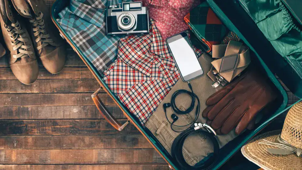 Open suitcase packed with clothes, gloves, a camera, a smartphone, and travel essentials on a wooden floor with boots and a straw hat beside it.