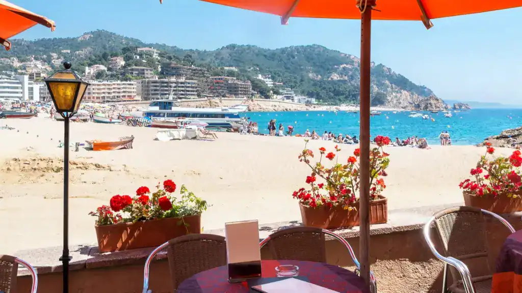Outdoor café view overlooking Tossa de Mar Beach in Costa Brava, with bright flowers, umbrellas, and turquoise water in the background.